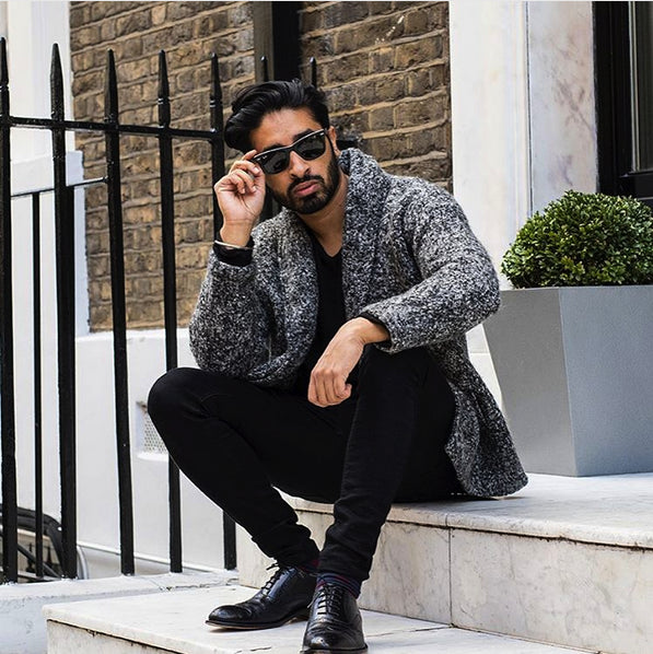 Man sitting on steps wearing a gray textured coat and black shoes, with a brick building and plant in the background and classic KOCH & CO. brogues